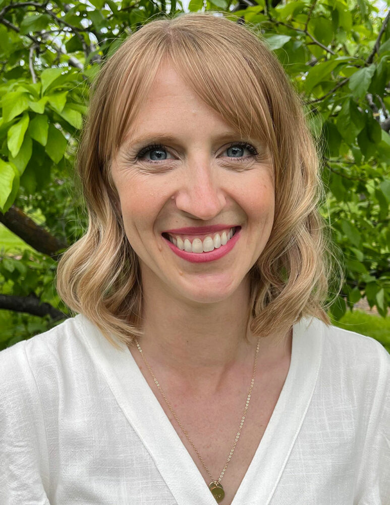 woman in white top smiles in front of tree