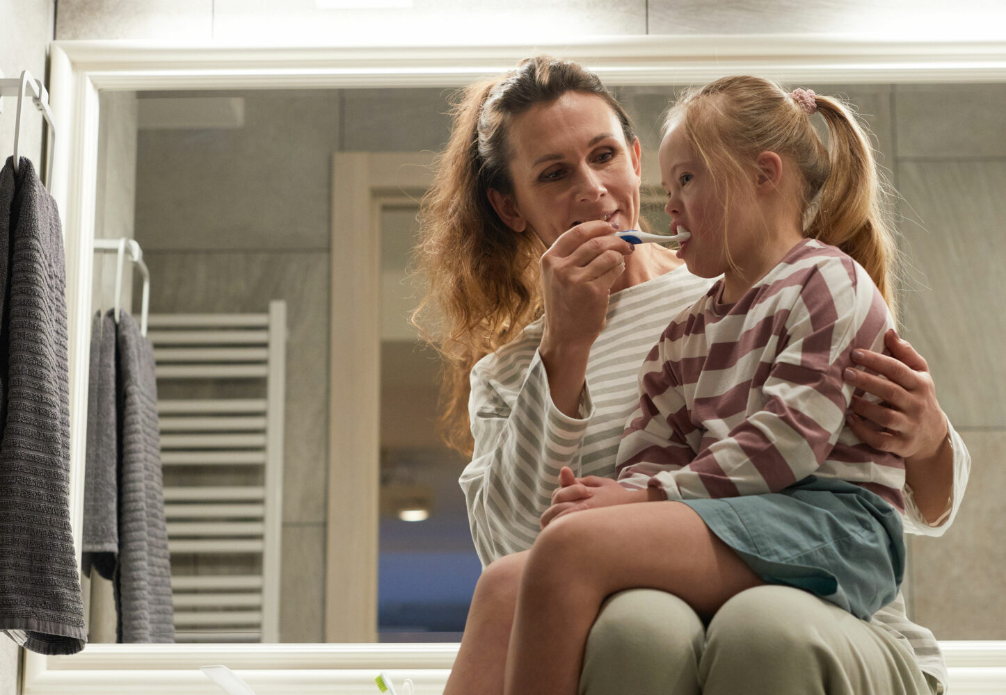 Mother brushes teeth of her daughter with Down syndrome who is sitting on her lap as mom sits on bathroom counter