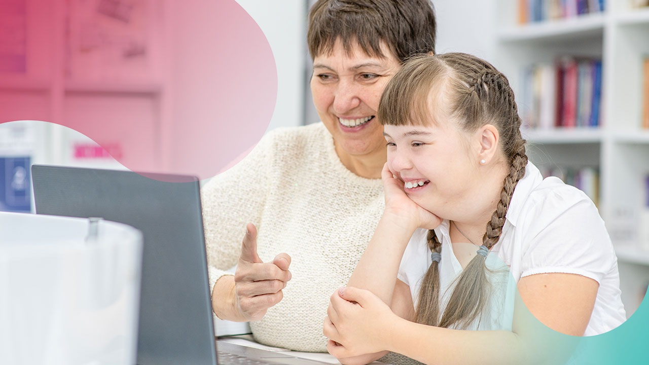 Female teenager with Down syndrome looks at laptop screen alongside female teacher in a library