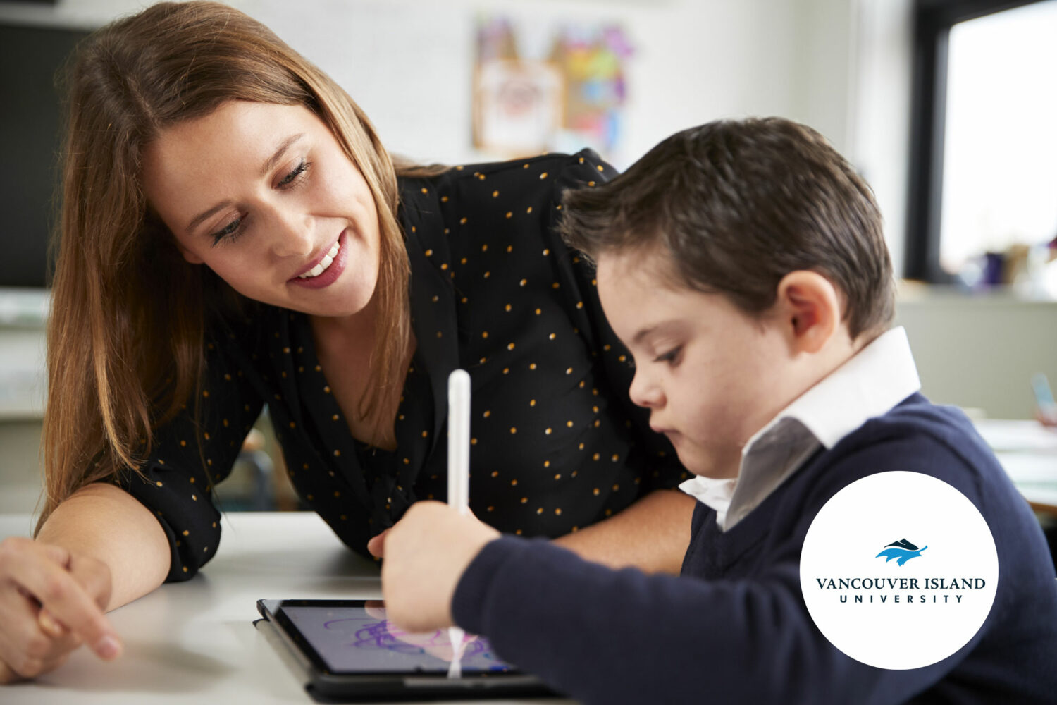 brown haired white boy with Down syndrome, wearing school uniform, writes on mobile tablet while female teacher looks on