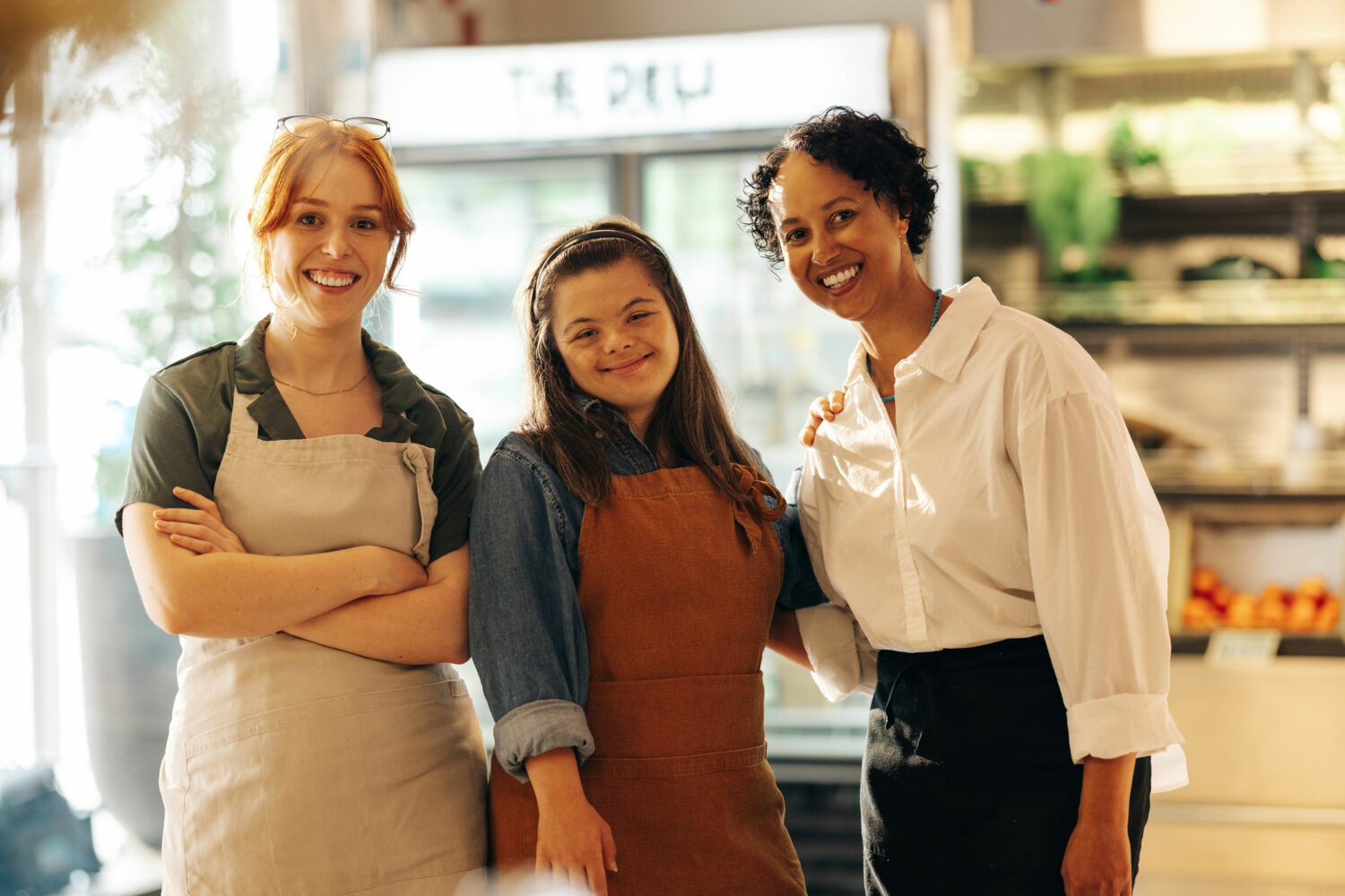 brown haired woman with down syndrome wearing denim shirt and brown apron, standing in grocery store with red headed white woman and black woman (co-workers)