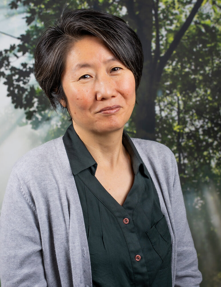 asian woman wearing green shirt and grey sweater in front of forest background