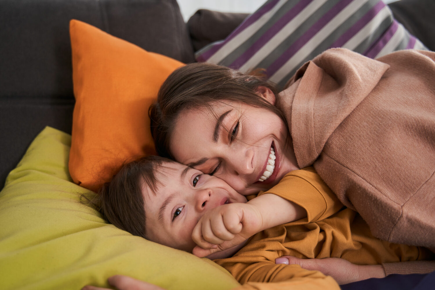 brown haired boy with Down syndrome lying on couch cushions, cuddling with brown haired mom