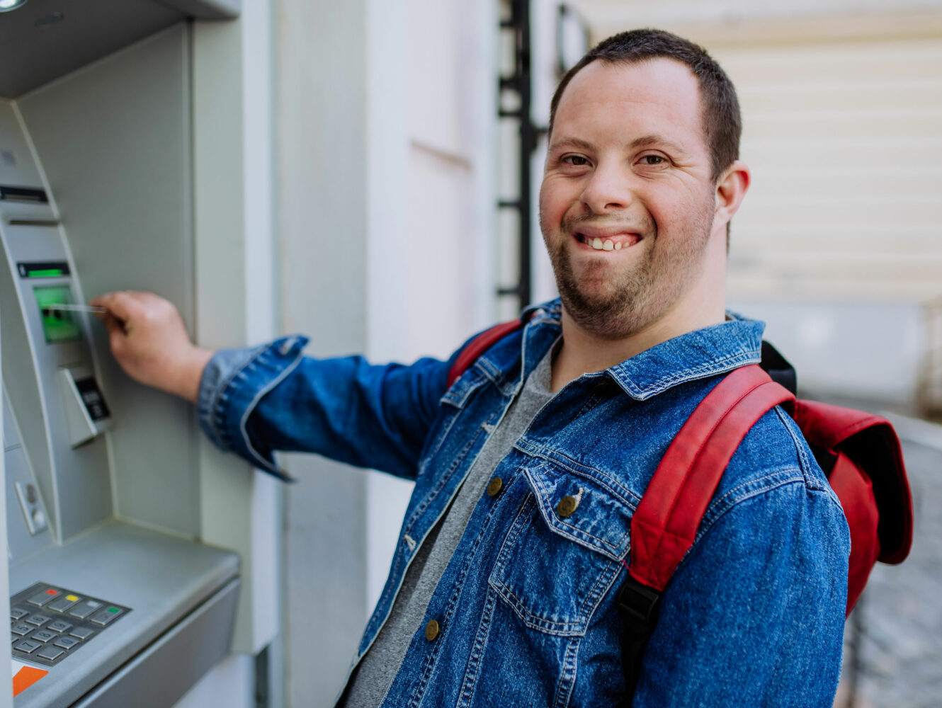 white man with Down syndrome wearing jean jacket and red backpack puts card in ATM
