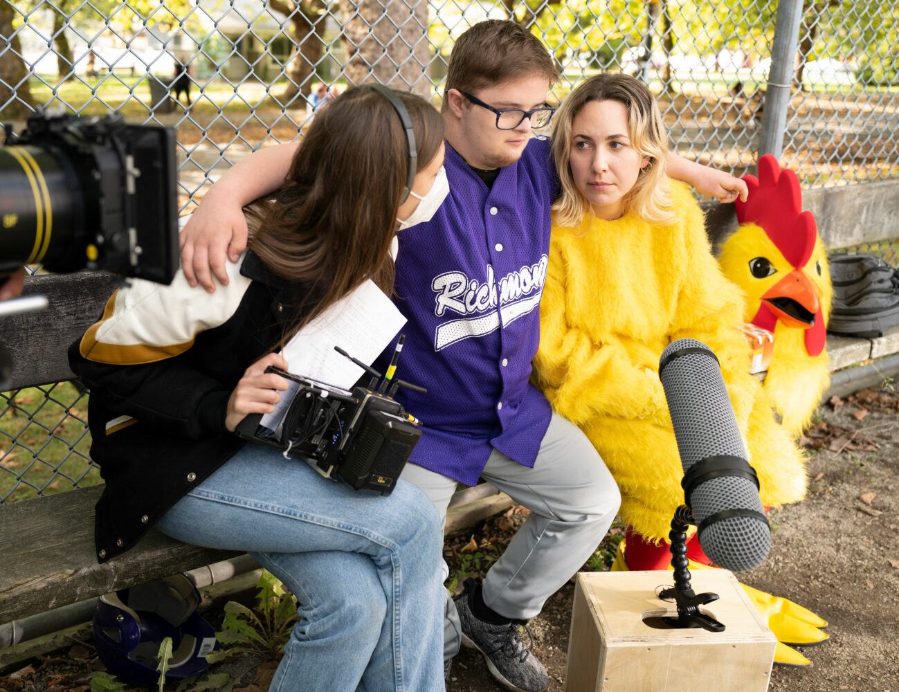 On a film set, young man with Down syndrome sits on bench wearing purple baseball jersey, with one arm around blonde woman in chicken costume and other arm around brown haired female film producer