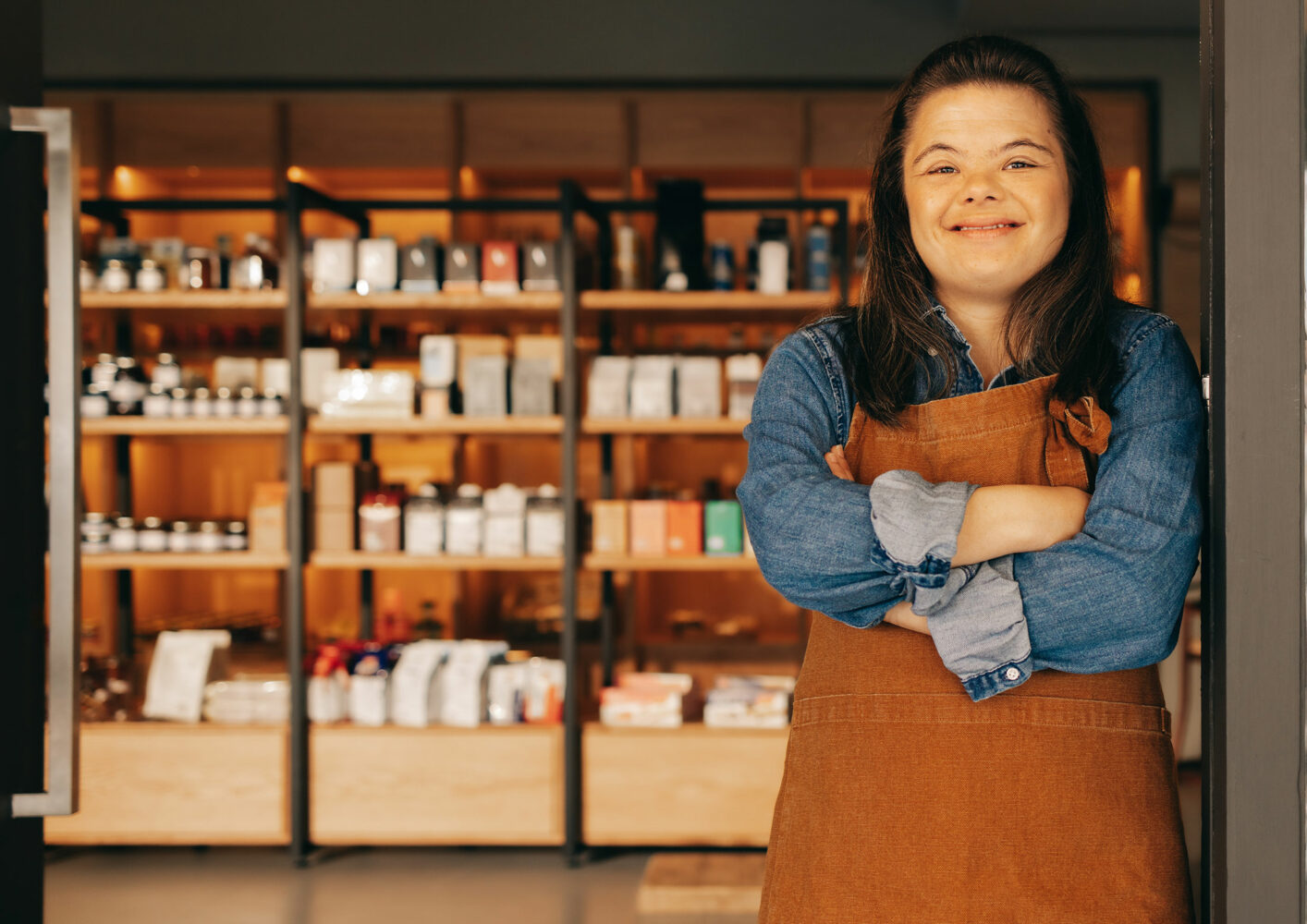 dark haired woman with Down syndrome wearing denim shirt and brown apron leans with arms crossed against doorway of the grocery store where she works