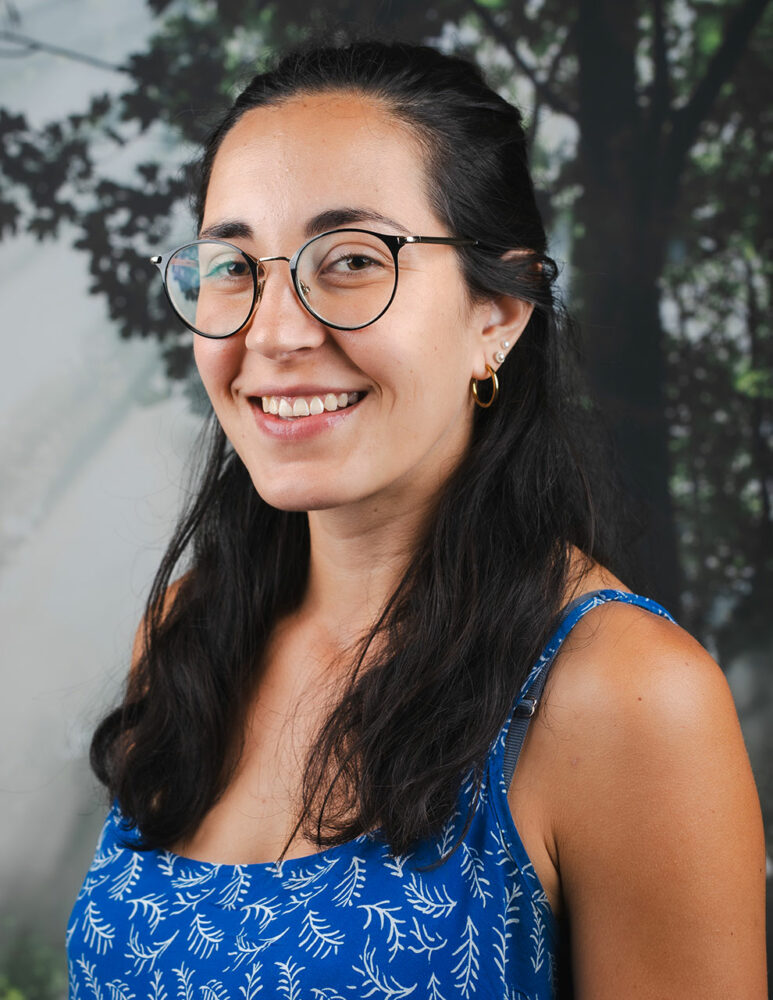 Latino adult female with long dark hair, glasses, and blue and white patterned dress in front of a picture of a tree