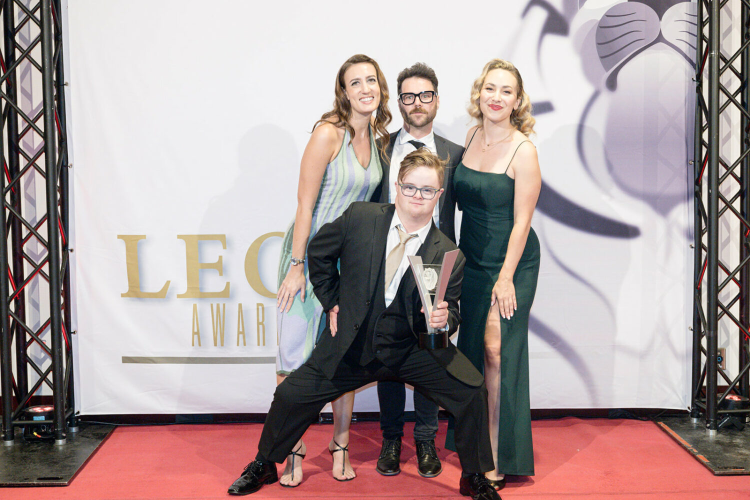 young man with Down syndrome, wearing glasses and a black suit, crouches down while holding a Leo award, with two white women and a white man wearing awards ceremony outfits behind him