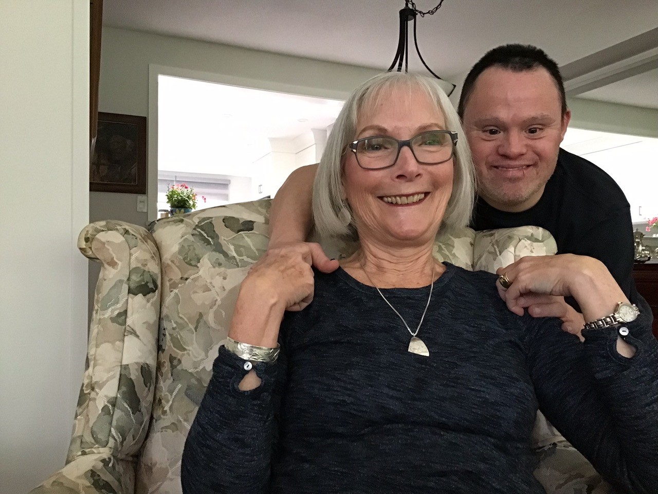 senior white white woman sitting on a chair, embraced from behind by her adult son with Down syndrome