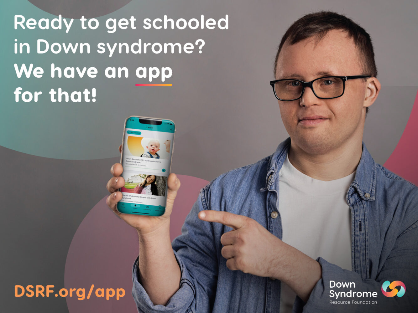 white man with Down syndrome, with dark hair, glasses, white t-shirt and blue long sleeve shirt, points at mobile phone displaying Down Syndrome Academy app