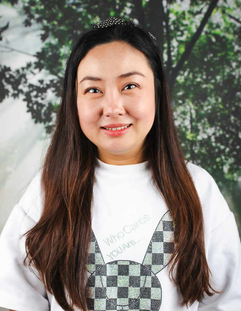 asian woman with long dark hair wearing white t-shirt smiling in front of picture of a tree