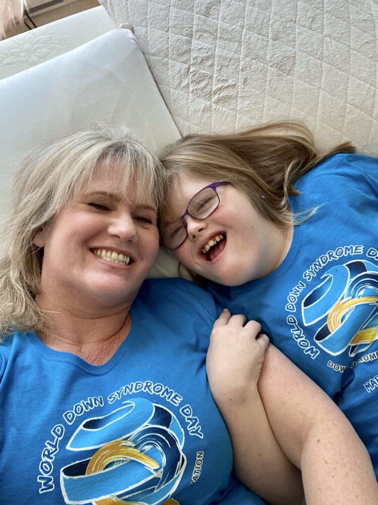 white female teenager with Down syndrome lies back on bed with her blonde-haired mother, both wearing turquoise t-shirts