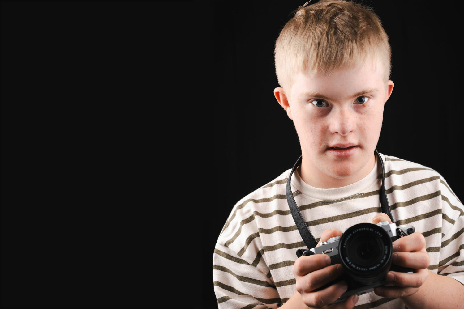 blonde teenage boy with Down syndrome wearing striped shirt with camera around his neck, on a black background
