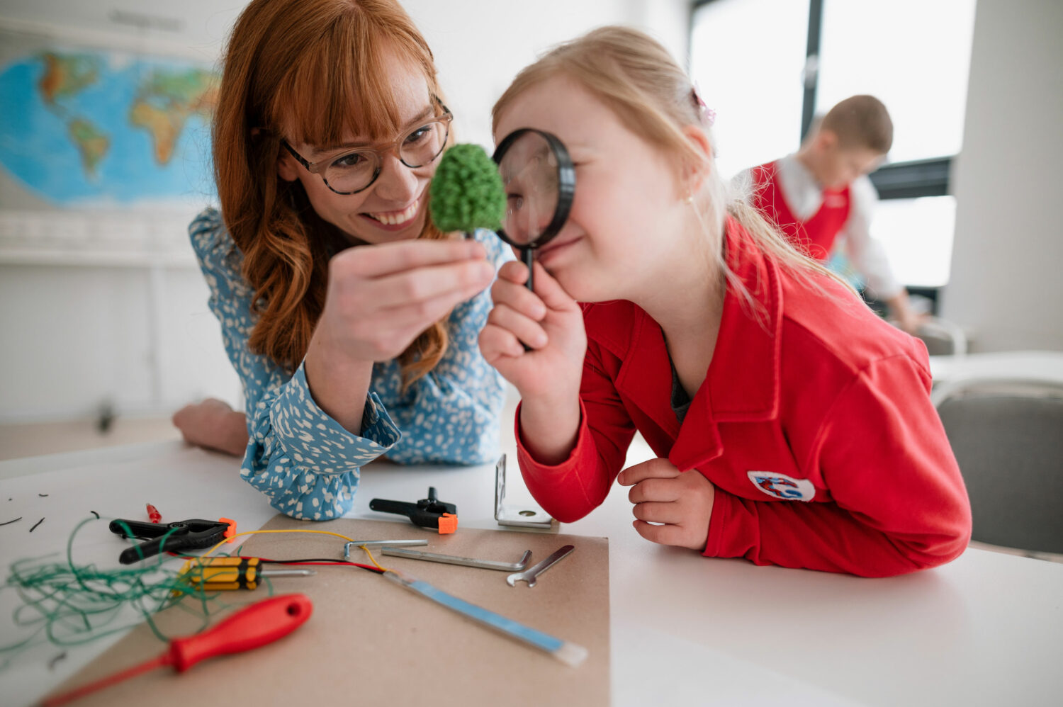 female student with Down syndrome wearing red sweater looks through magnifying glass with assistance from her female teacher