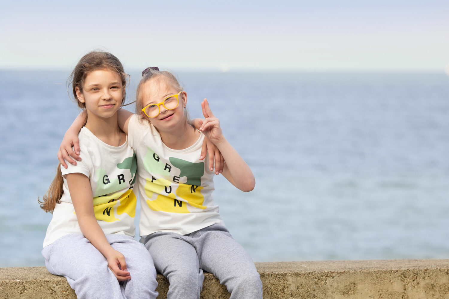 2 girls, one with Down syndrome, sit together with ocean behind them, with arms around one another