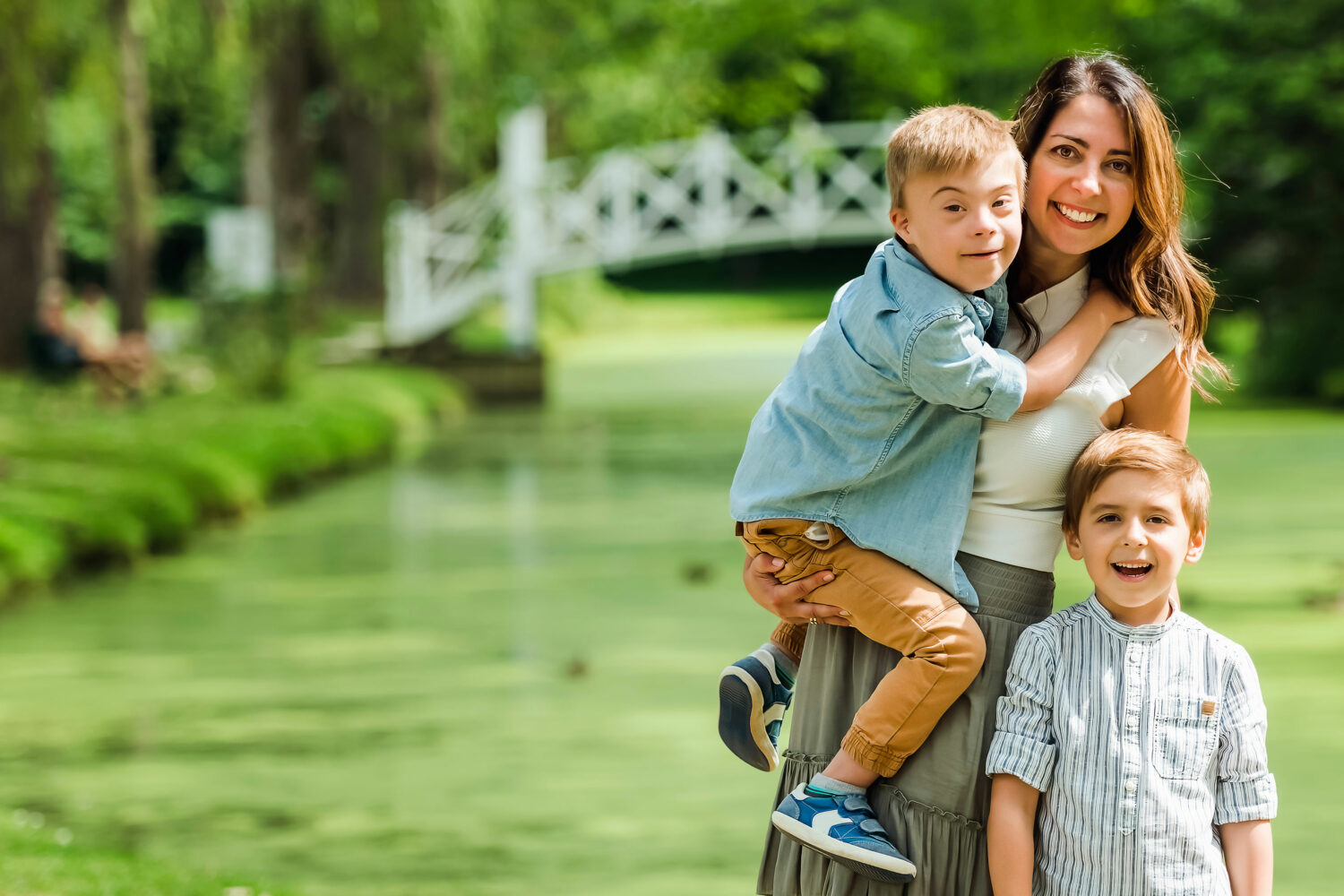 white mom with long brown hair holds her young son with Down syndrome on her hip with his older brother standing beside her, in front of outdoor greenery and white bridge