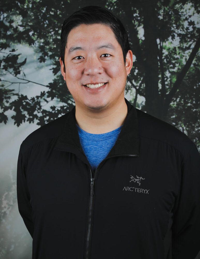 dark haired Asian man wearing black jacket over blue t-shirt smiles in front of picture of a tree