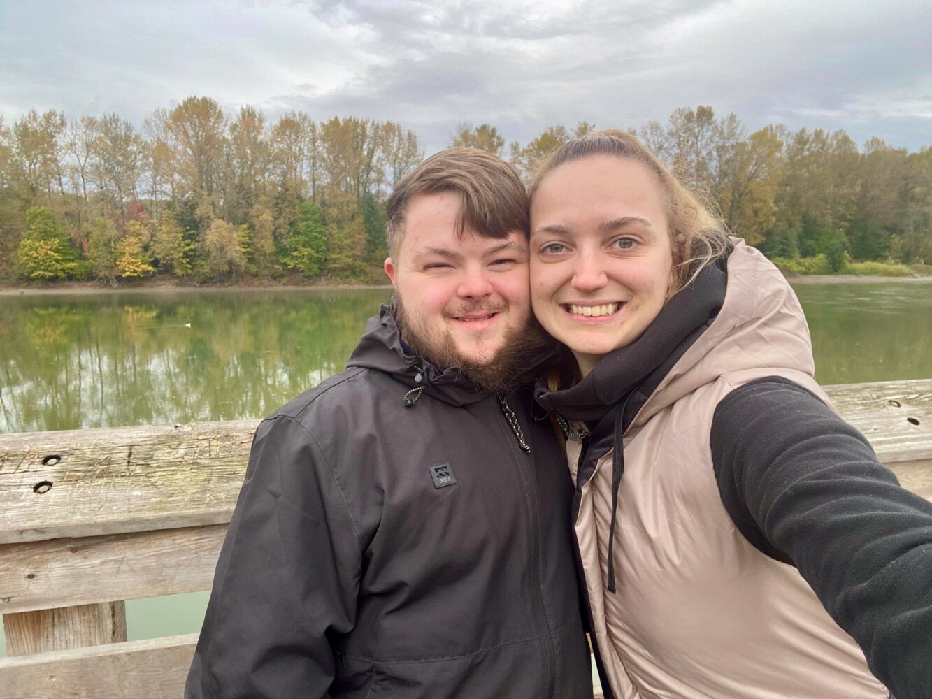 2 young adult siblings: a white man with Down syndrome and his older sister, posing together with a river behind them
