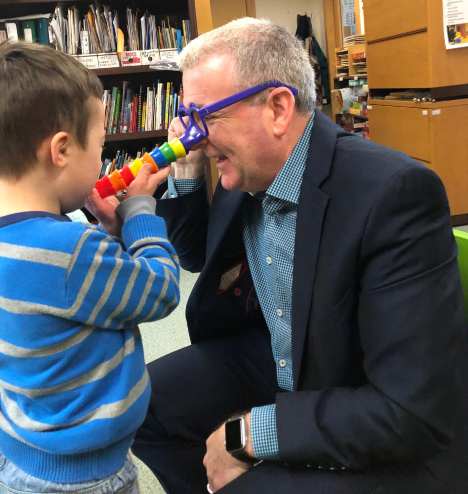 white man in a business suit sits on the floor and plays with young boy with Down syndrome, who puts a toy tube over the man's nose