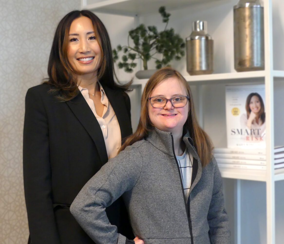 a white woman with Down syndrome with long brown hair, glasses, and gray sweater stand in front of Asian business woman