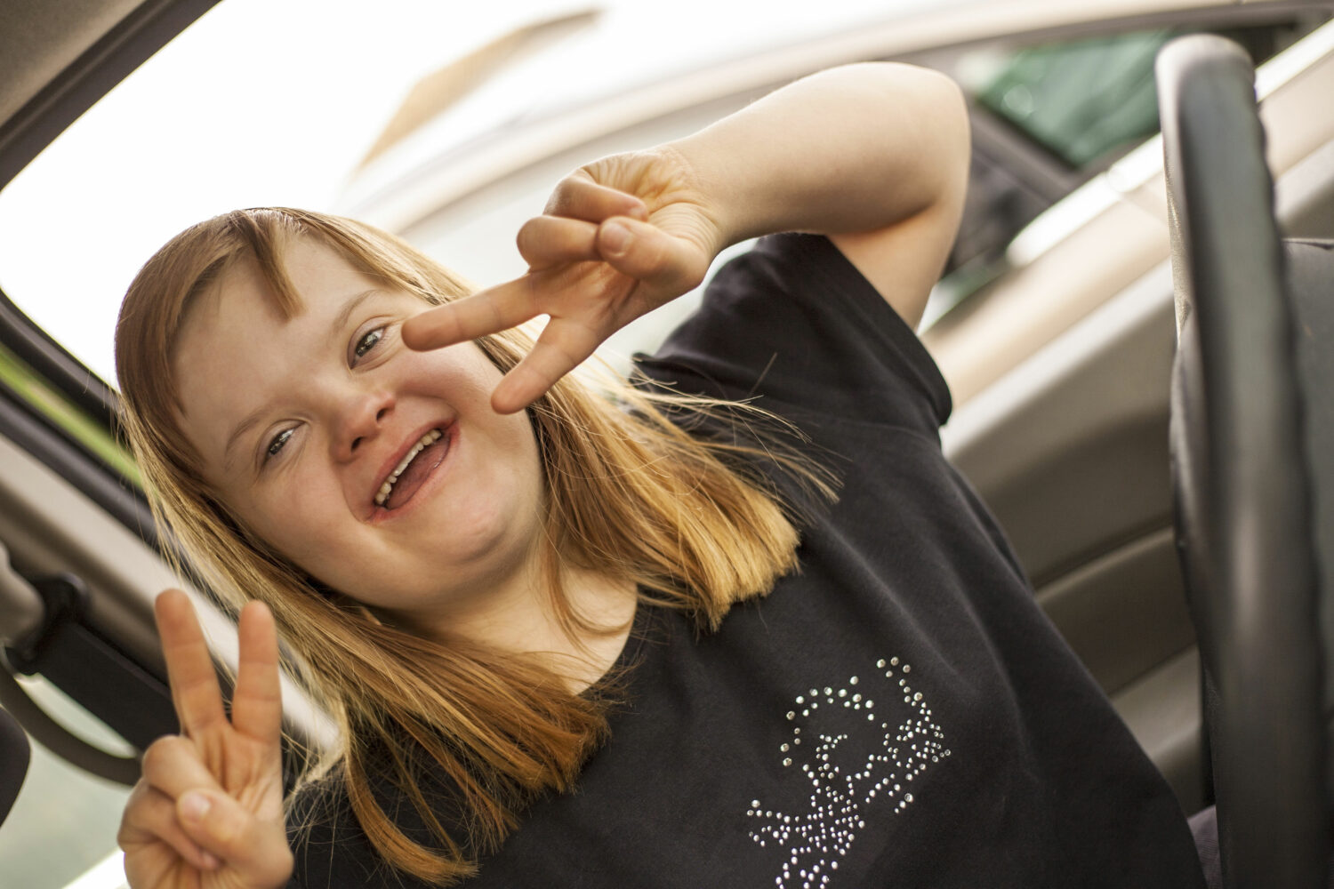 white woman with Down syndrome, with long blonde hair, sits behind the steering wheel of a car and flashes a double peace sign