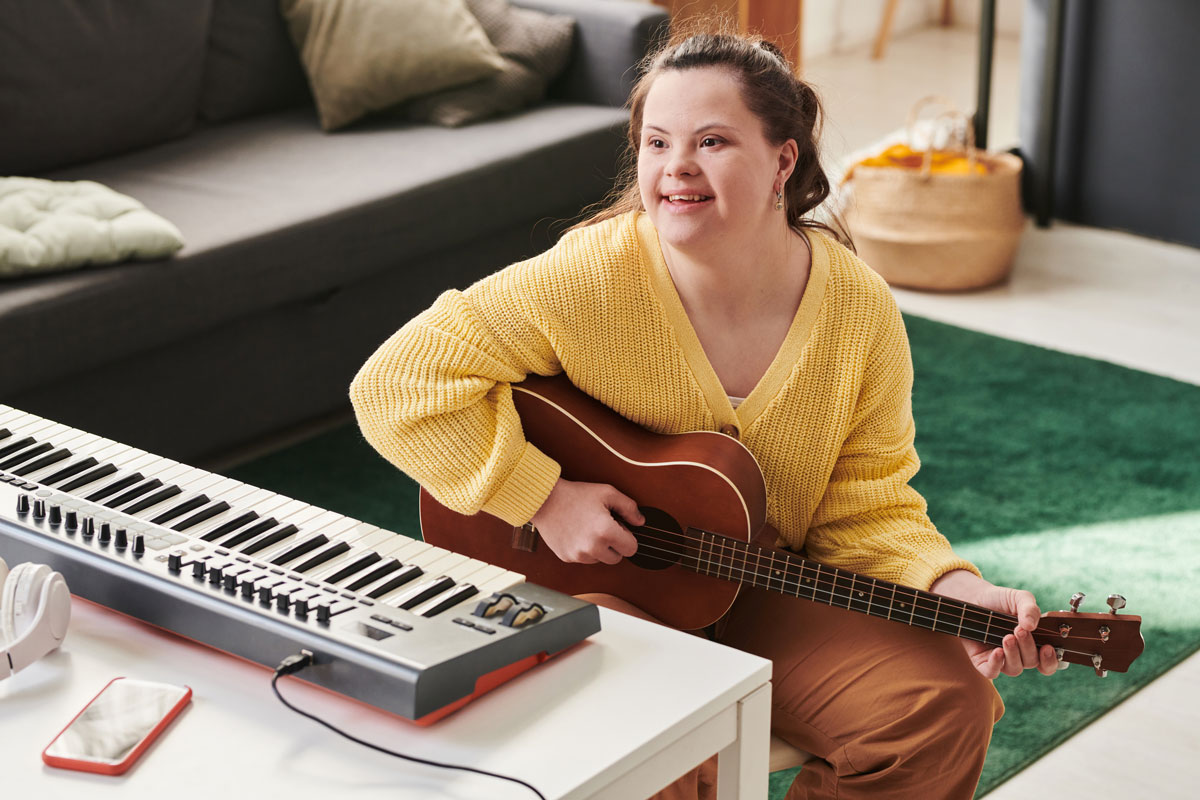 white woman with Down syndrome sits on floor and strums guitar with keyboard beside her