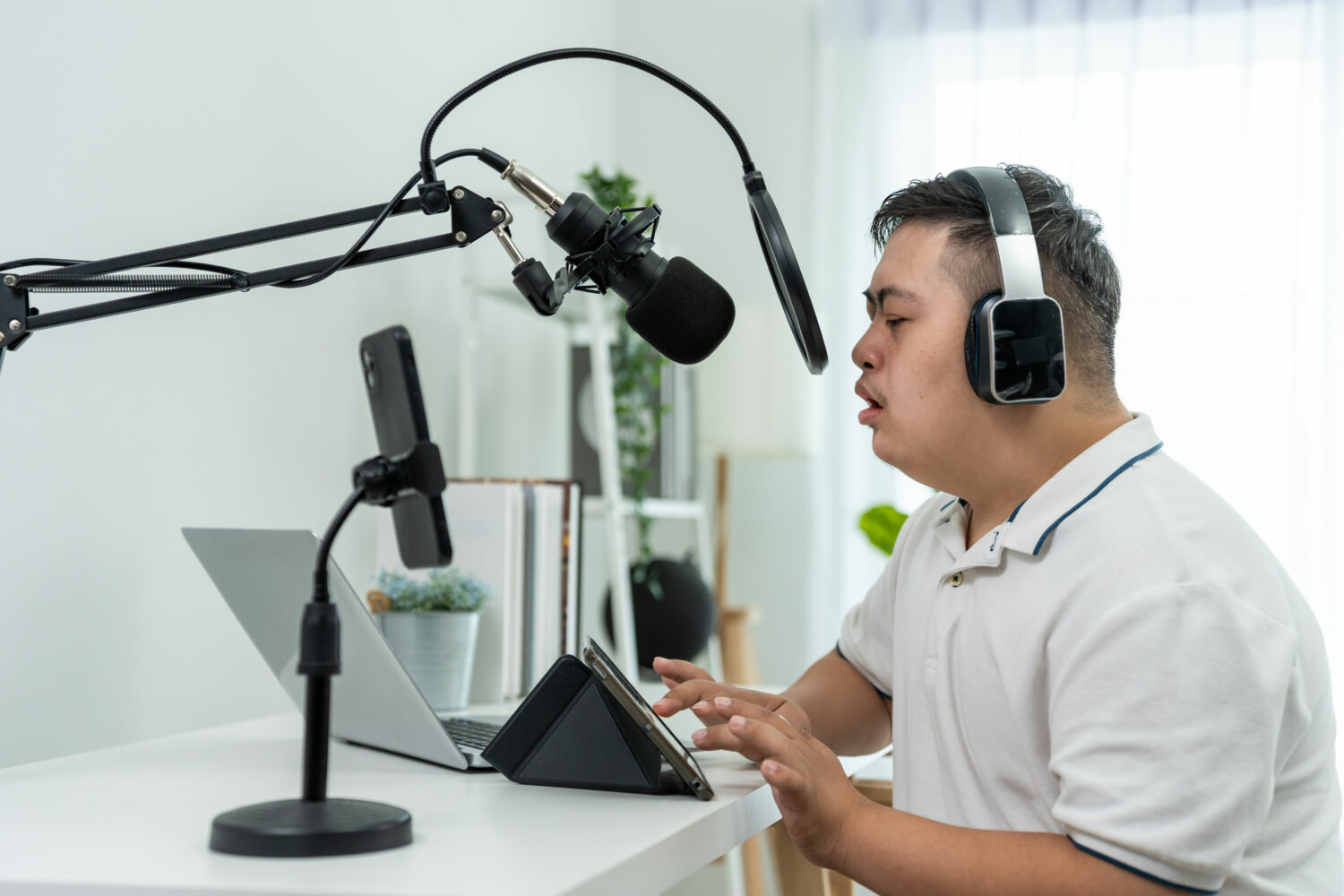 Asian man with down syndrome wearing headphones and white shirt sits at desk and speaks into microphone