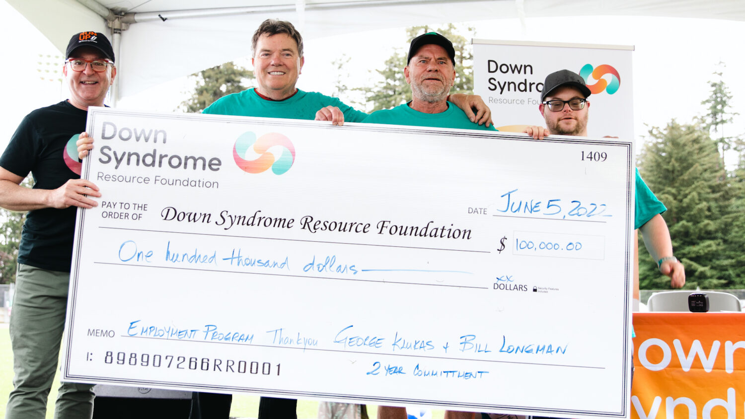 4 men, one of whom has Down syndrome, hold a giant novelty cheque for $100,000 made out to the Down Syndrome Resource Foundation
