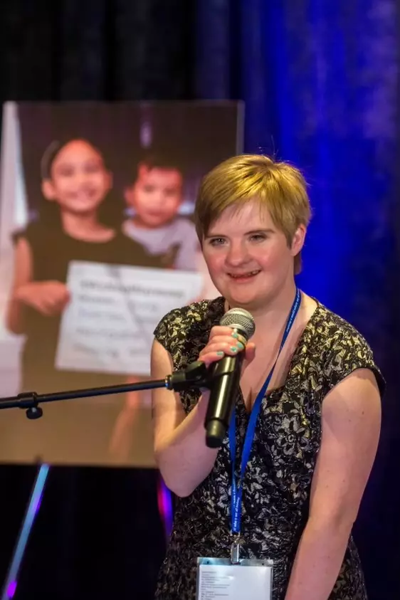 white woman with Down syndrome, with short blonde hair and black and gold top, speaks into a microphone