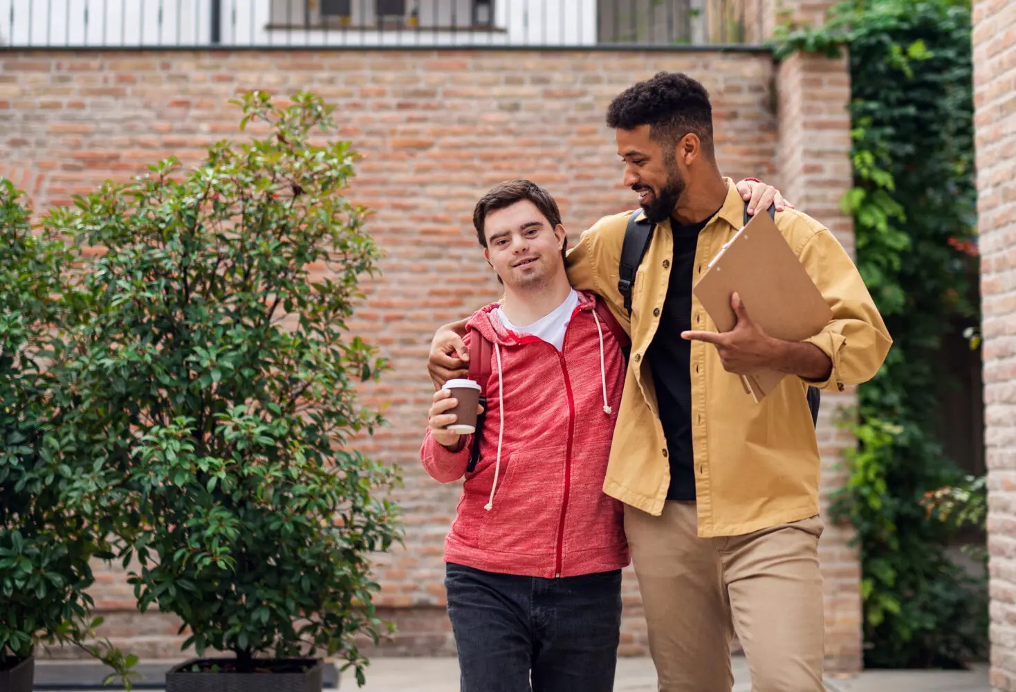 young white man with Down syndrome, with black pants and salmon sweater, walks with arm around taller black man wearing a tan jacket and beige pants, outdoors