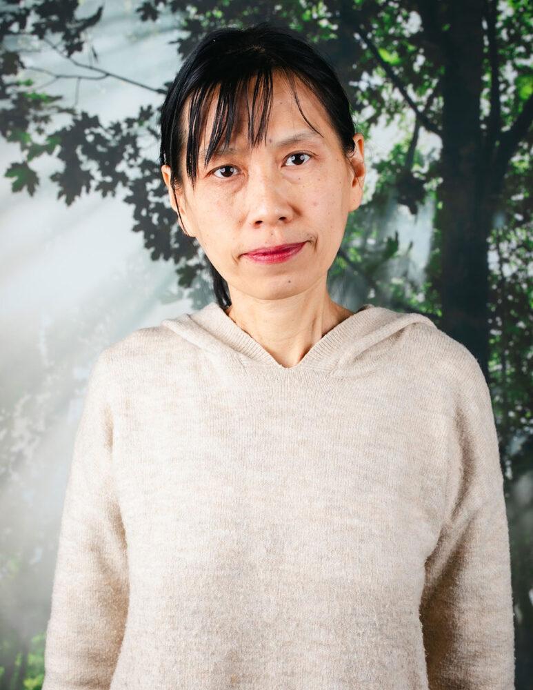 Asian woman with dark hair wearing beige sweater stands in front of a picture of a tree