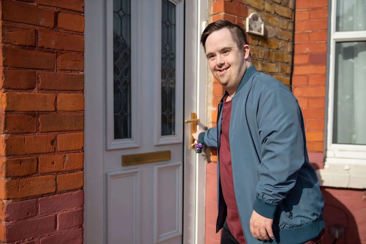 brown haired man with Down syndrome wearing blue jacket over red shirt opens the door to a brick house