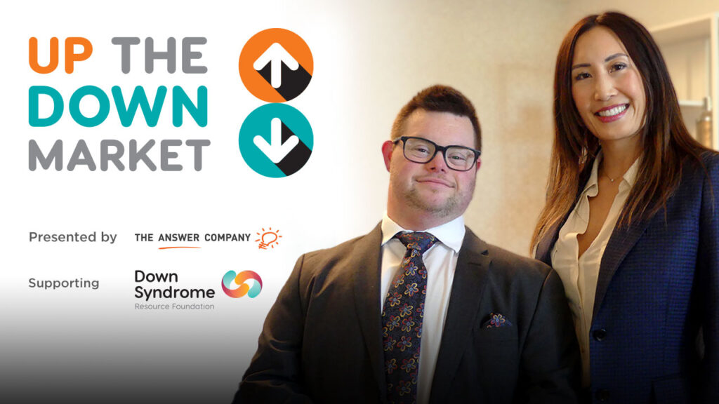 white, brown haired man with Down syndrome wearing a dark suit, next to a long-haired Asian woman wearing a white blouse and blue jacket, with logo for Up the Down Market, presented by The Answer Company and benefiting the Down Syndrome Resource Foundation
