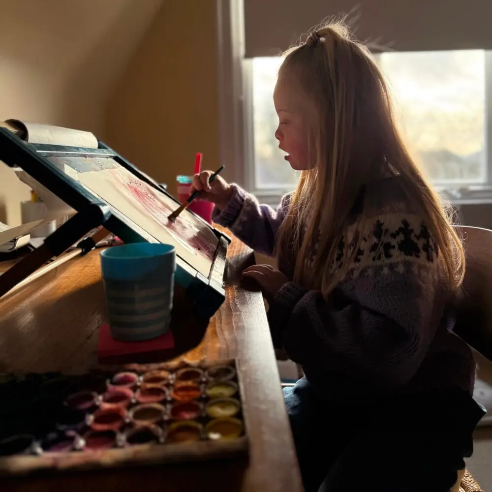 Image of a little girl with Down syndrome sitting at a desk painting with watercolors. She has a piece of paper on a slant board in front of her, with a palate of water colour paint and a cup of water to her left. She is sitting in profile, wearing a purple knit sweater, with long blonde hair. She is holding a paint brush to the paper in her right hand.