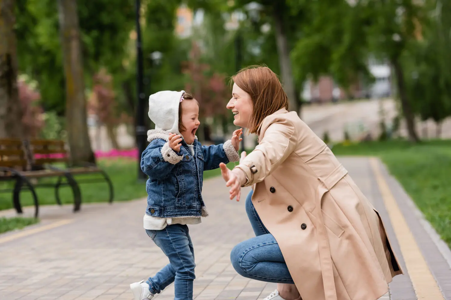 young girl with Down syndrome wearing blue denim pants and jacket and grey hood runs to embrace her mom, squatting down, wearing long beige coat and blue jeans, outdoors at a park