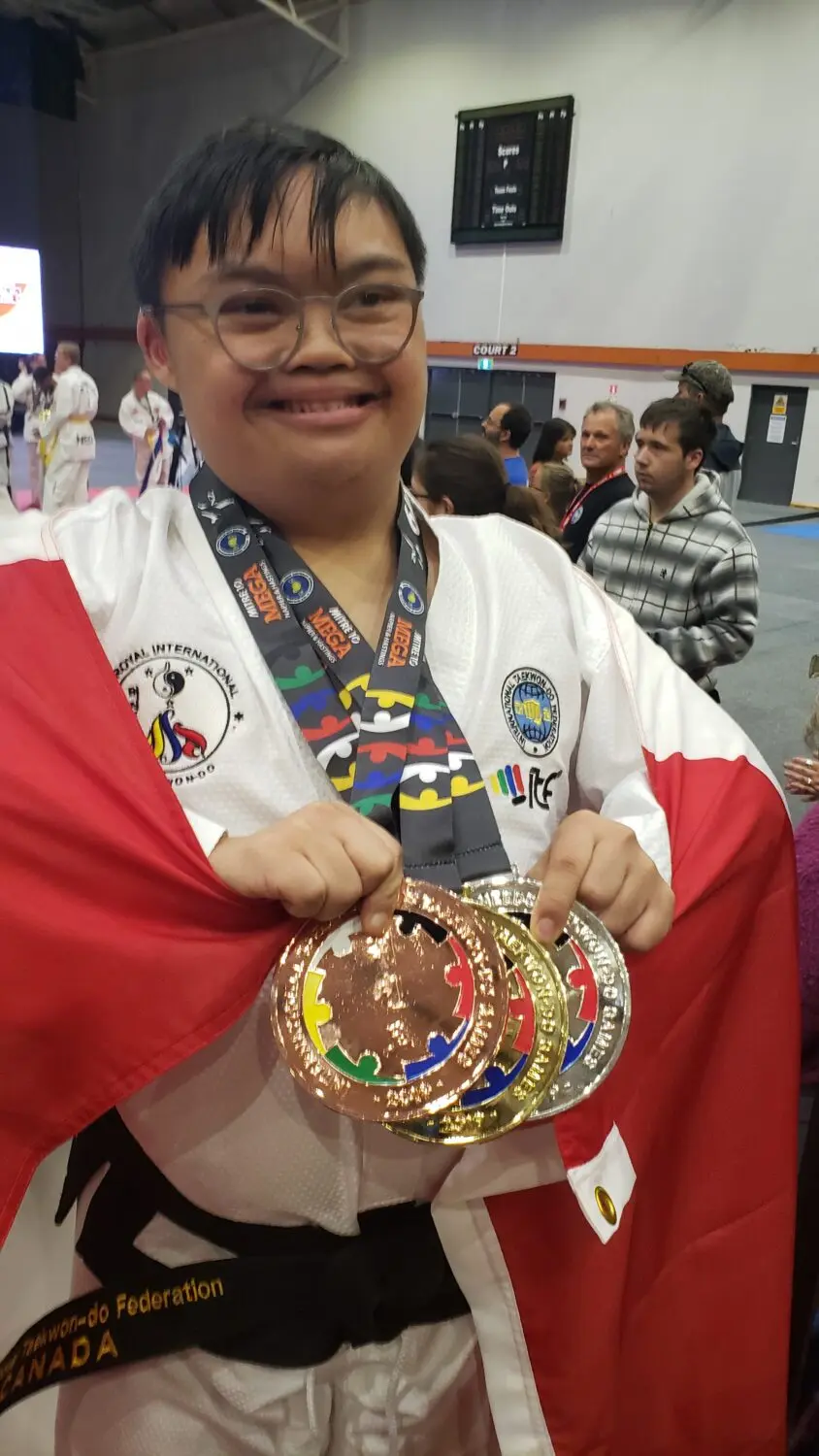 Asian man with Down syndrome wearing a white taekwondo uniform with a Canadian flag draped around his shoulders and three large medals around his neck