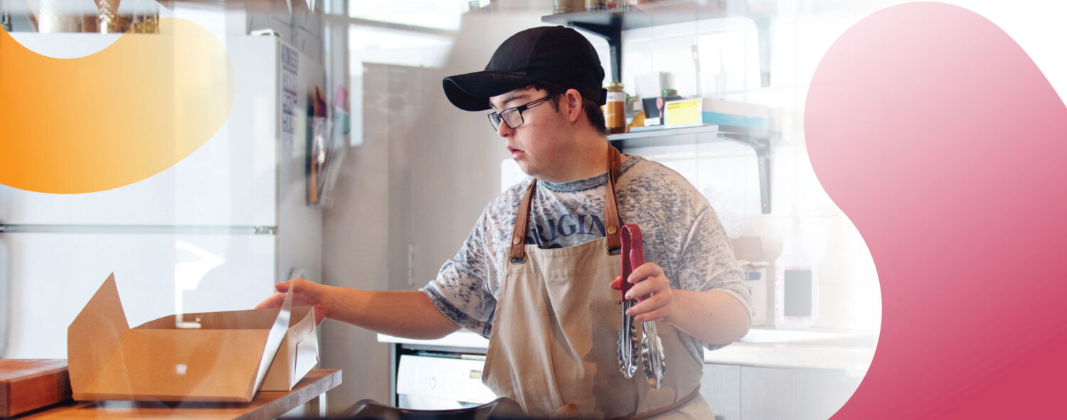 a young man with Down syndrome working in a commercial kitchen, wearing a black cap and tan apron, with a food box in one hand and a pair of tongs in the other