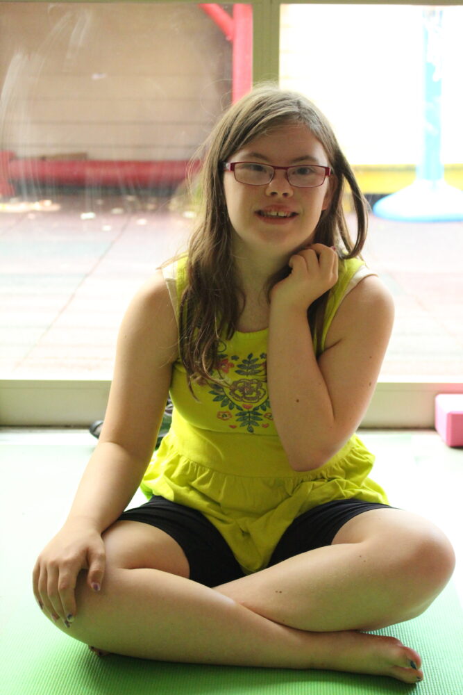 young woman with Down syndrome, with long brown hair and glasses, wearing a yellow tank top and black shorts, sits cross legged on a green yoga mat